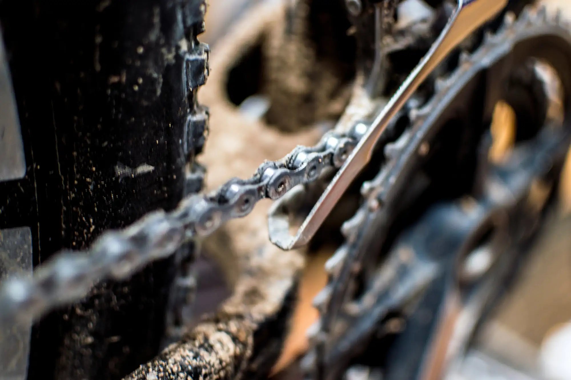 Cadena de bicicleta negra cubierta de suciedad con eslabones metálicos y una herramienta plateada descansando sobre ella.