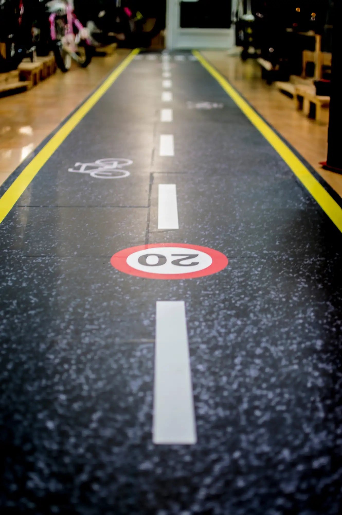 Suelo negro moteado con marcas de carril bici pintadas en blanco y amarillo, incluyendo una señal de límite de velocidad de ’20’’ km/h circular roja.